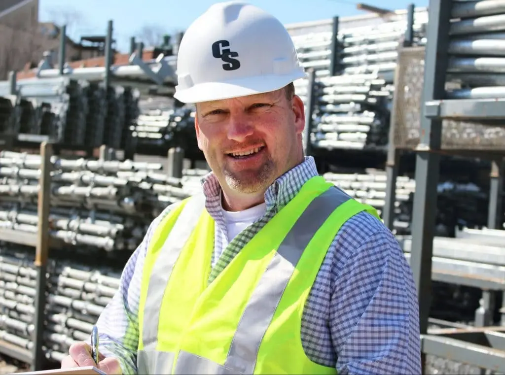 A smiling engineer wearing safety gear and taking notes at a construction site.